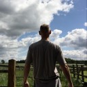 A man with his back to us, looking into the distance and a blue sky with a few light clouds.