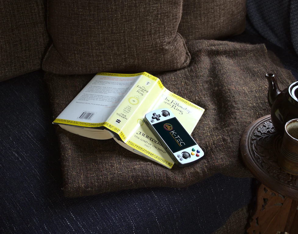 Grey Anbernic RG405M retro gaming handheld resting on a classic book with a teapot, representing a relaxing, ready-to-play gaming experience.