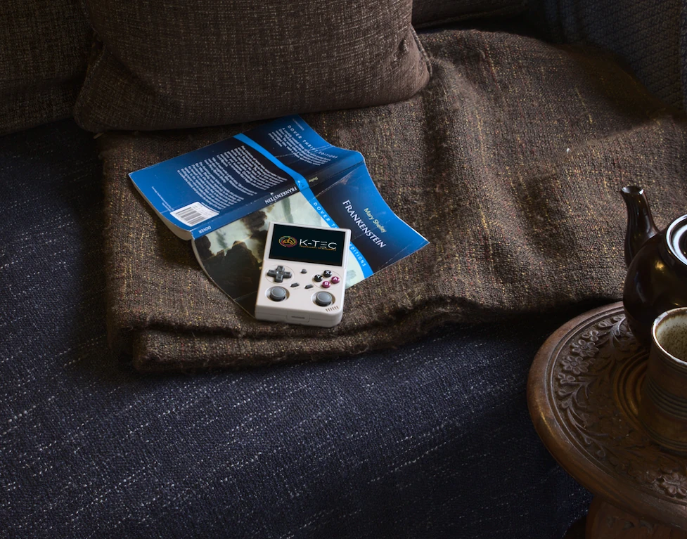 BATLEXP G350 in Retro Grey resting on a copy of Mary Shelley's Frankenstein on a cosy sofa with a teapot and mug nearby — a relaxing afternoon of retro gaming and classic British literature